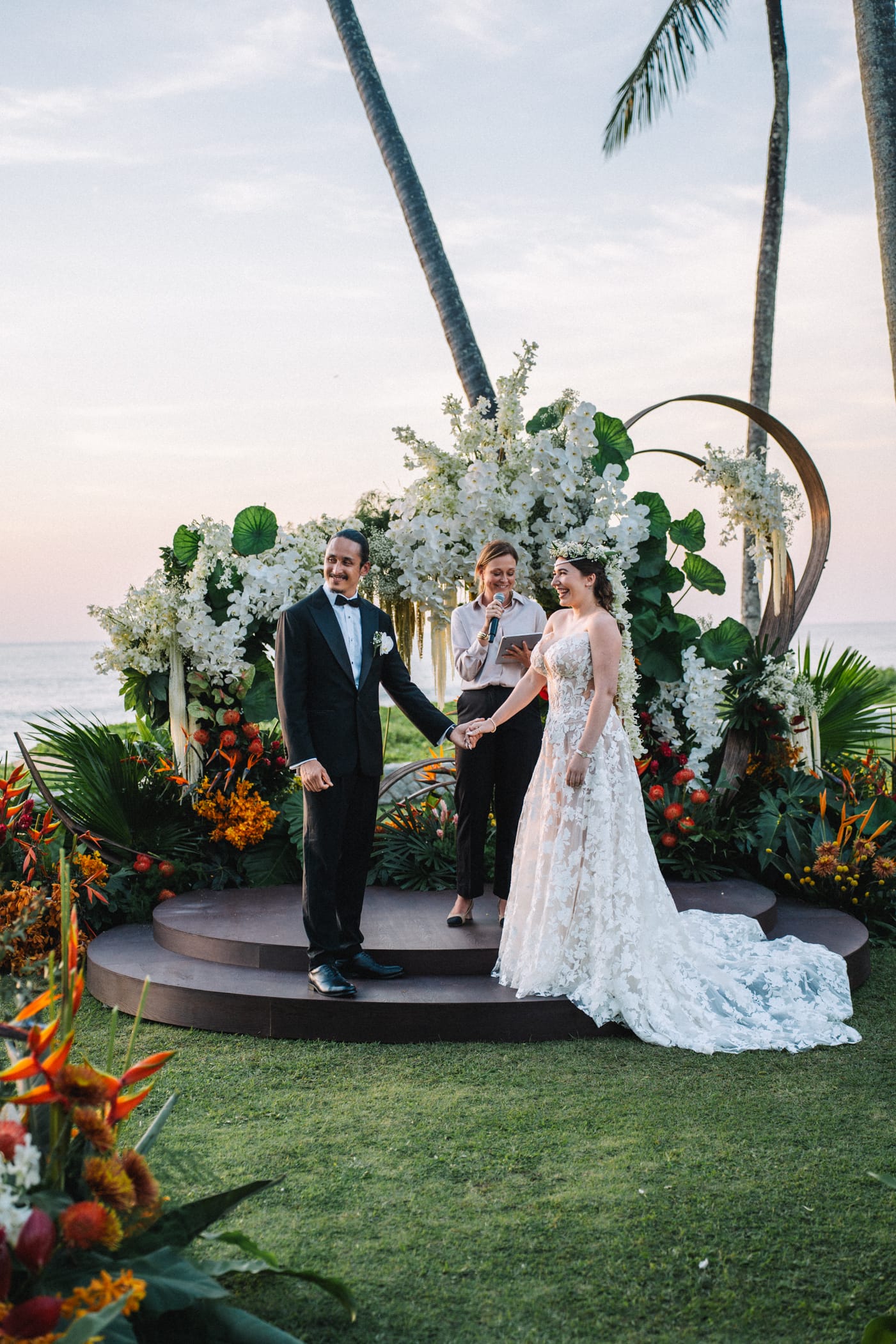 Bride and groom standing under floral wedding arch facing the sea during ceremony